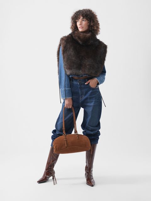 Eine Person, die eine Jeansjacke, dunkle Jeans und einen dunklen Pelzschal trägt, eine braune Handtasche aus Wildleder in der Hand hält und hohe braune Stiefel aus geprägtem Leder trägt.
