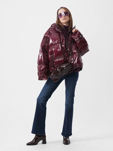 Ein Model, das eine glänzende, tiefrote Daunenjacke, eine dazu passende Umhängetasche, dunkelblaue Jeans und eine getönte Sonnenbrille trägt.