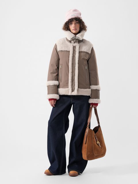 Ein Model, das eine hellbraune Jacke mit cremefarbenem Kunstfellbesatz, eine blaue Jeans mit weitem Bein und eine rosa Mütze trägt und eine braune Schultertasche aus Wildleder in der Hand hält.