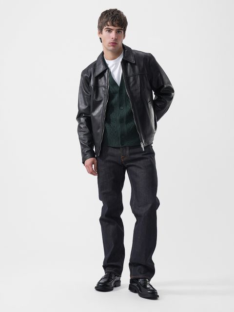 Ein Mann, der eine schwarze Lederjacke über einer grünen Strickjacke, einem weißen T-Shirt, einer dunklen Jeans und schwarzen Slippern trägt.