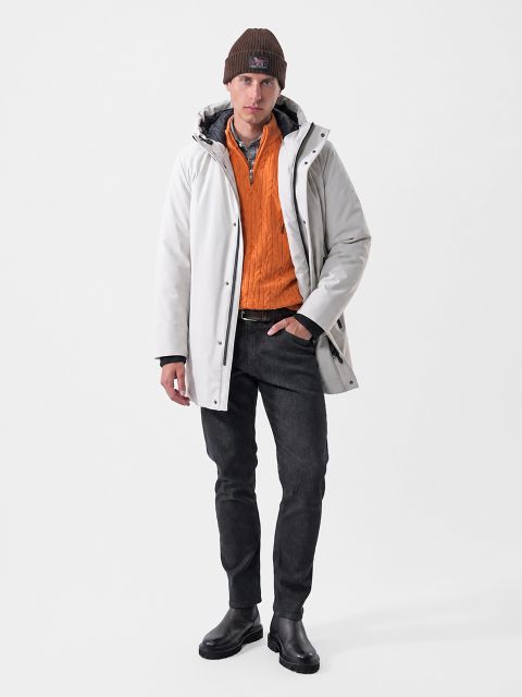 Ein Mann, der einen weißen Parka mit Kapuze, einen orangefarbenen Pullover mit Viertelreißverschluss, schwarze Jeans und eine braune Mütze trägt.