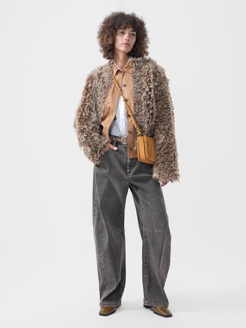 Eine Person, die eine Jacke mit flauschiger Struktur über einem hellbraunen Hemd mit weißem Kragen trägt, dazu eine graue Jeans mit weitem Bein und eine hellbraune Umhängetasche.