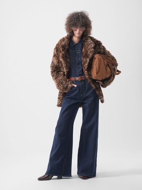 Eine Person, die einen braunen Pelzmantel über einem dunklen Jeans-Overall mit braunem Gürtel trägt und eine braune Wildledertasche in der Hand hält.