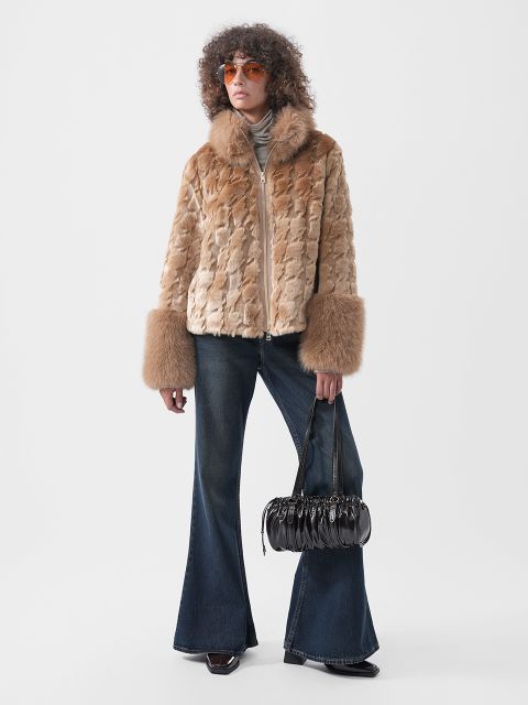 Eine Person, die eine hellbraune Kunstfelljacke mit breiten Ärmelbündchen, eine dunkle Schlagjeans und eine orangefarbene Sonnenbrille trägt und eine schwarze, plissierte Handtasche in der Hand hält.