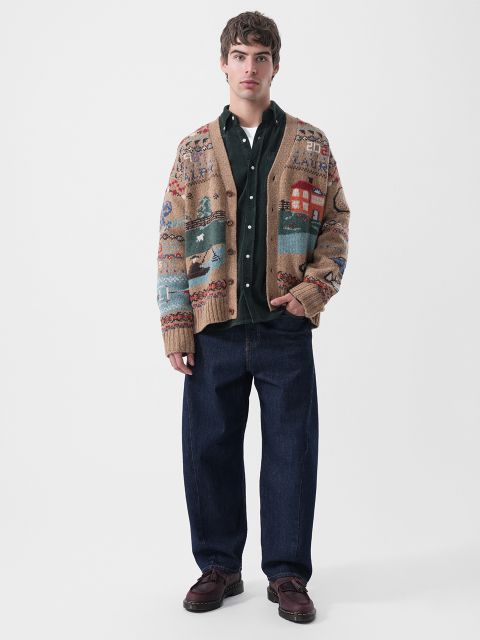 Ein Mann, der eine hellbraune, gemusterte Strickjacke, ein dunkelgrünes Hemd mit Knöpfen und eine dunkelblaue Jeans trägt.