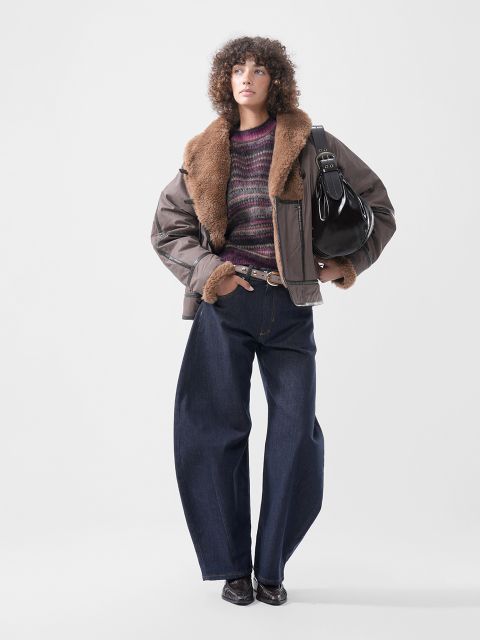 Eine Person, die eine braune Jacke mit pelzbesetztem Kragen, einen lila gestreiften Pullover und eine dunkle Jeans mit weitem Bein trägt und eine schwarze Umhängetasche bei sich hat.