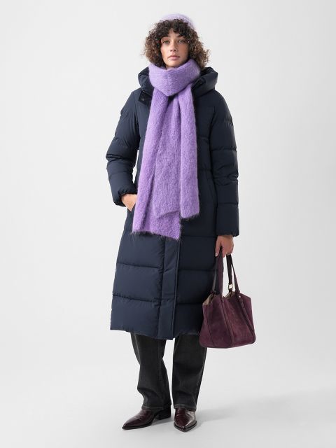 Eine Person, die einen langen marineblauen Daunenmantel, eine lila Mütze und einen dazu passenden lila Schal trägt und eine dunkelrote Handtasche in der Hand hält.