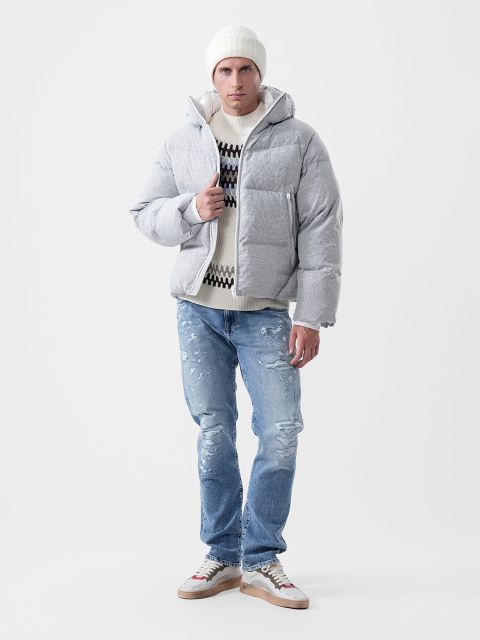 Ein Mann, der eine hellgraue Daunenjacke, einen gemusterten Pullover, eine weiße Mütze und ausgewaschene blaue Jeans sowie Turnschuhe trägt.
