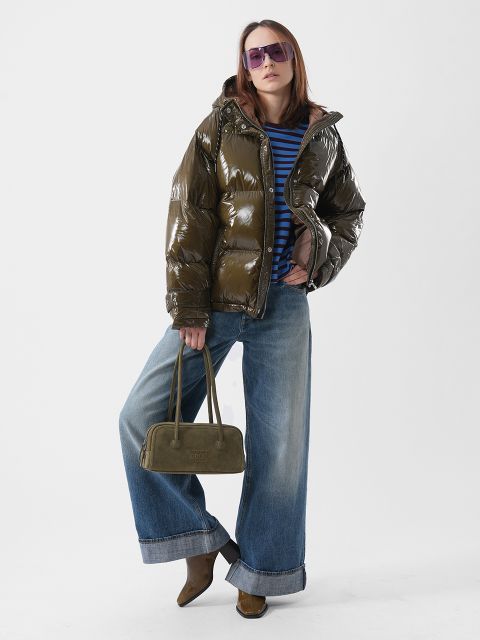Eine Person, die eine glänzende, olivgrüne Daunenjacke, ein blau gestreiftes Hemd, eine Jeans mit weitem Bein und eine lila Sonnenbrille trägt und eine olivgrüne Handtasche in der Hand hält.