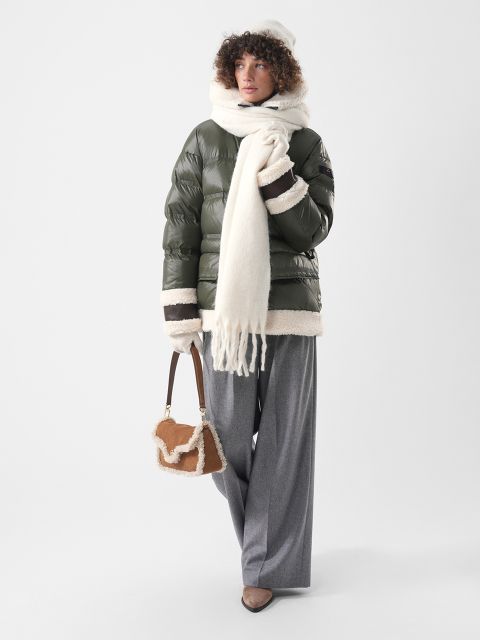 Eine Person, die eine olivgrüne Daunenjacke mit Lammfellbesatz, einen weißen Schal und eine Mütze sowie eine graue Hose trägt und eine kleine braune Tasche mit Lammfellbesatz bei sich hat.